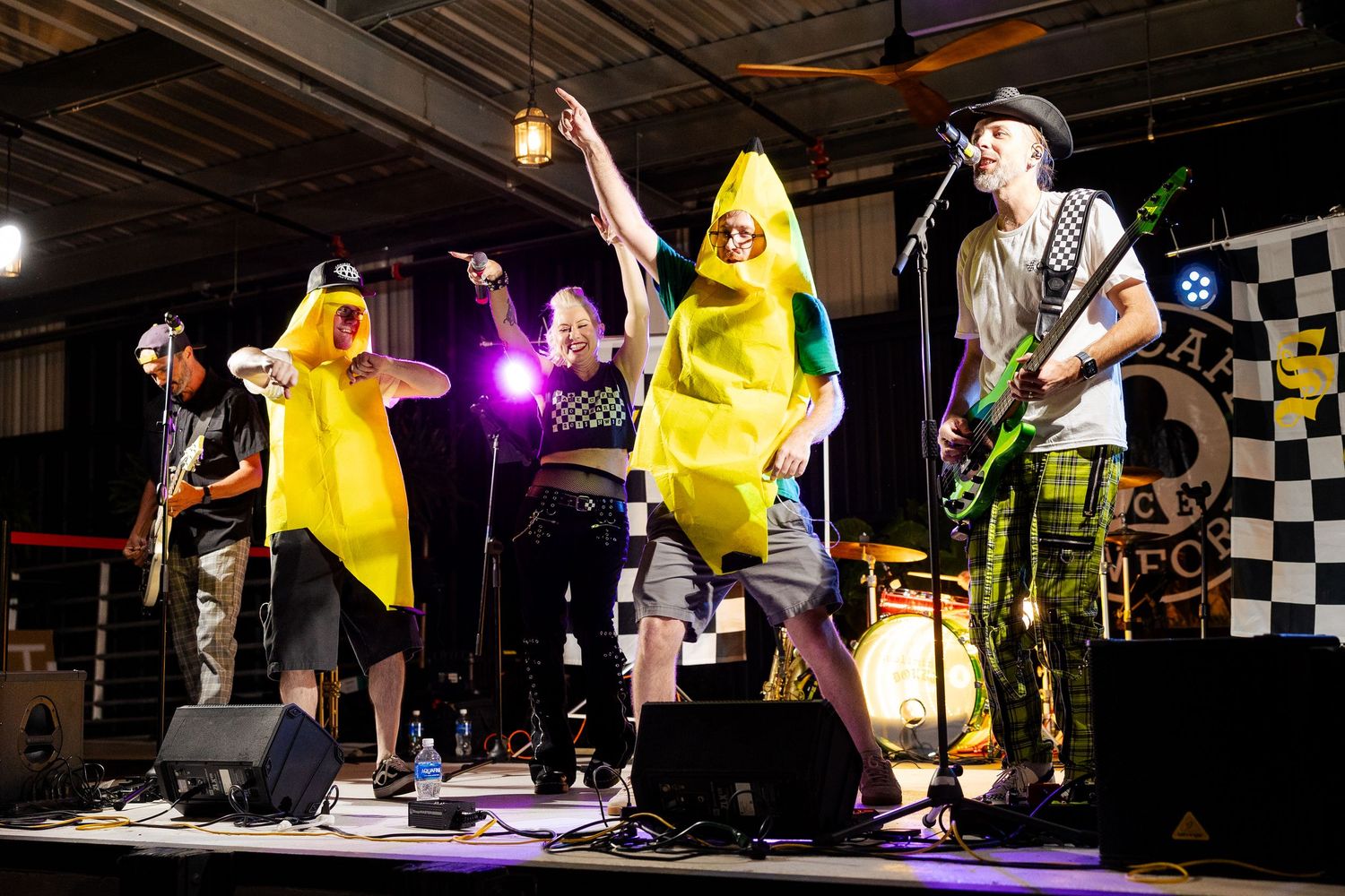 Band performing in banana costumes on stage with guitarist in checkered pants at live music event.