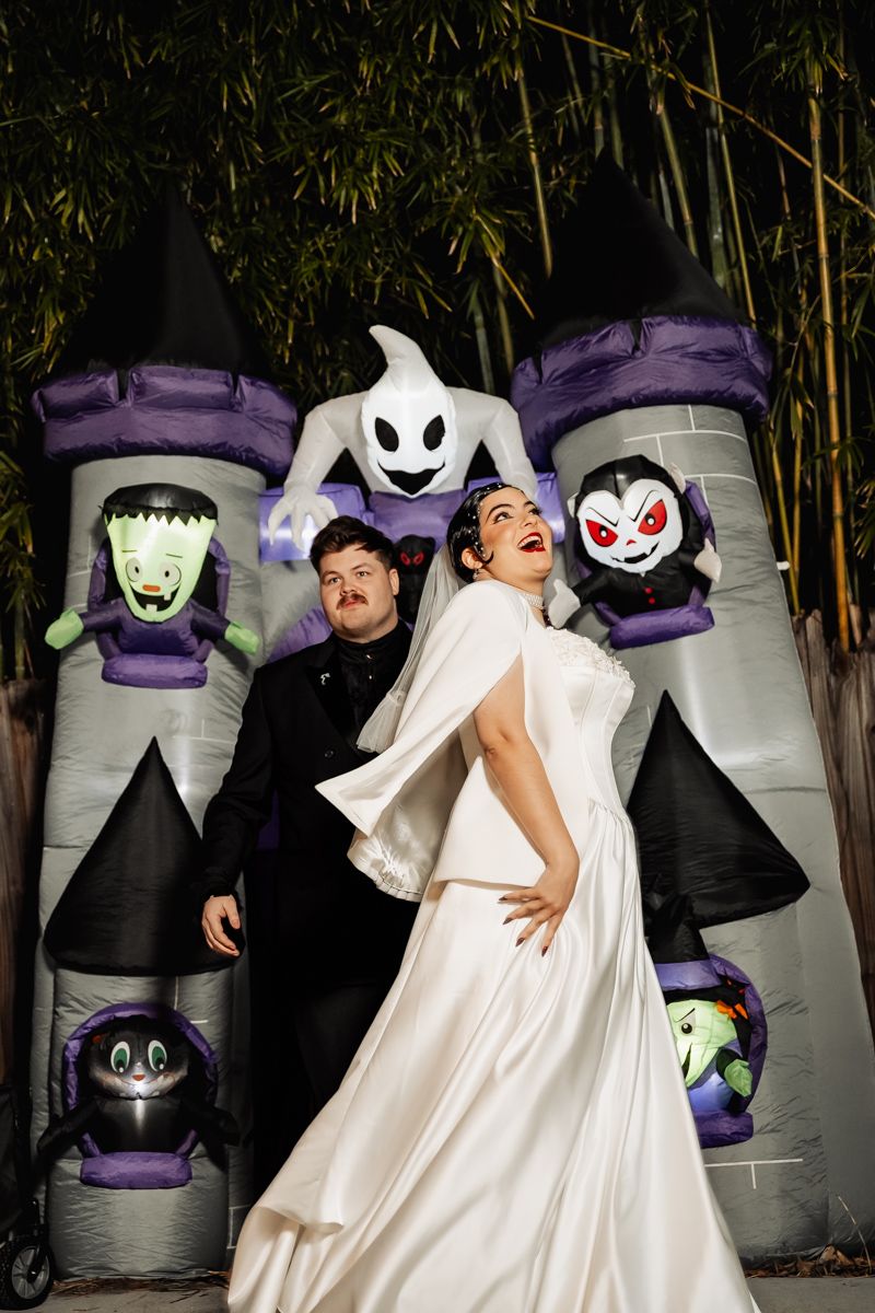 Bride and groom pose playfully in front of inflatable Halloween castle with ghost and monster decorations.