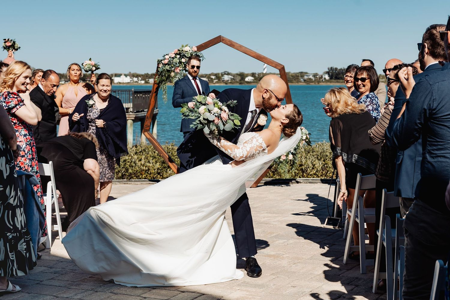 Groom dips bride in romantic kiss while guests cheer along the aisle at outdoor waterfront ceremony.