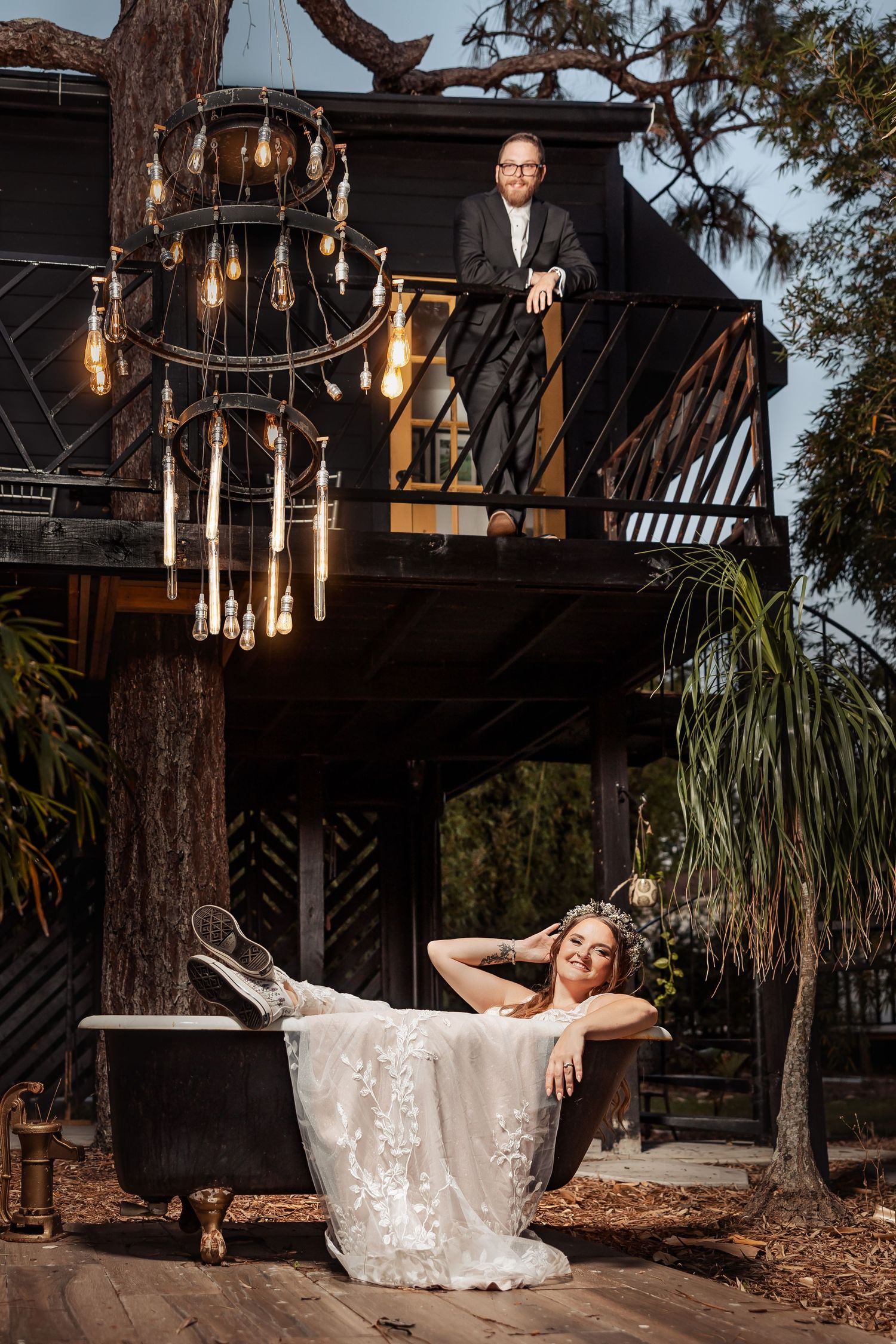 Elegant wedding photo with bride in vintage bathtub and vintage chandelier hanging from rustic treehouse balcony at sunset.