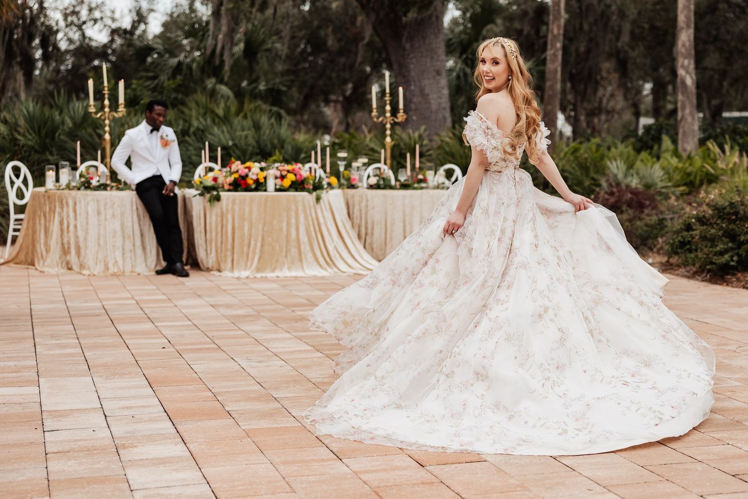 Bride in lace ballgown poses elegantly on outdoor terrace with reception tables and candle decor in background.
