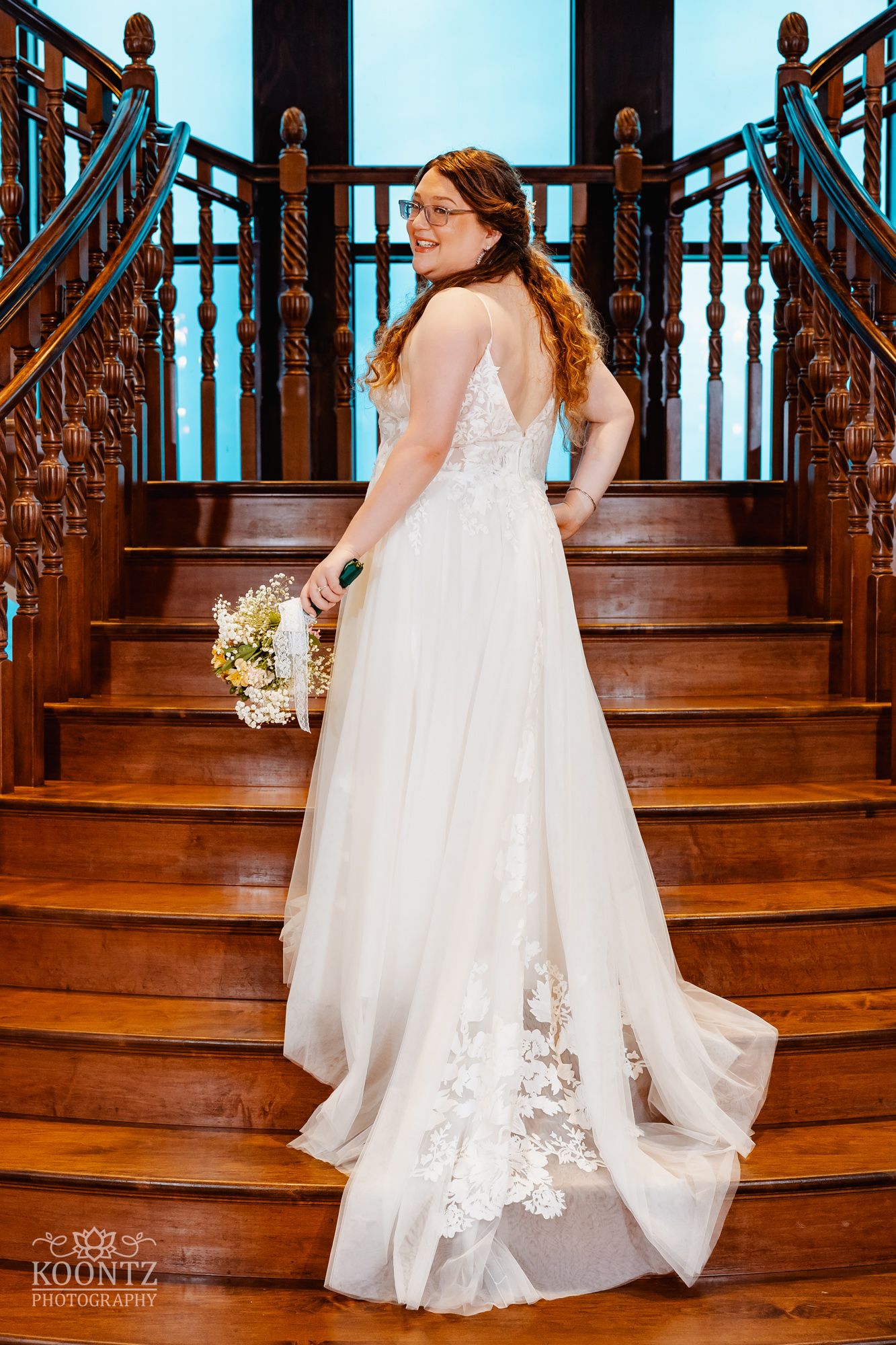 Bride in flowing white dress with lace details poses on grand wooden staircase.