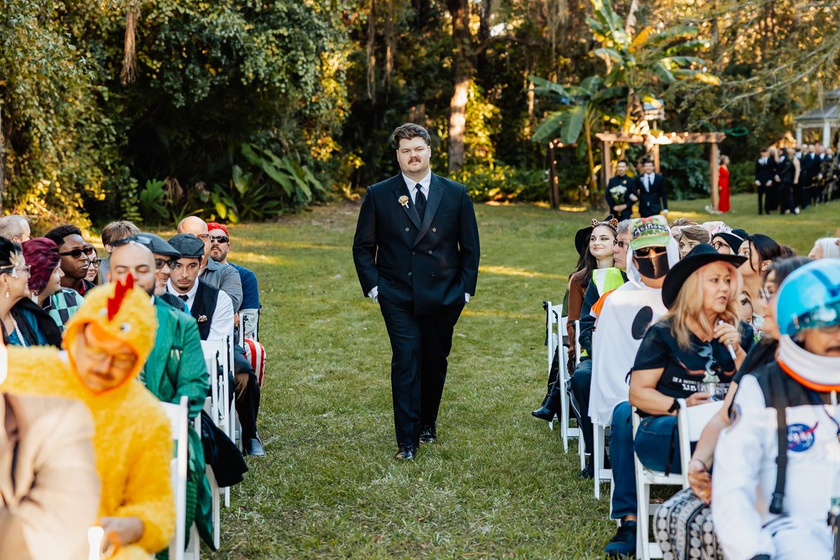 Guests seated in white chairs on a lawn during an outdoor wedding ceremony with autumn trees in the background.