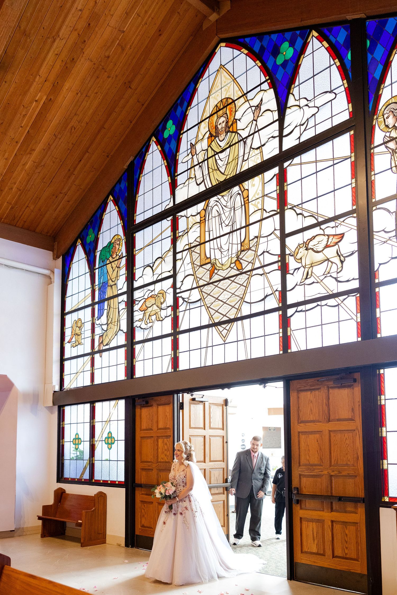 Bride and escort stand at church entrance beneath stunning stained glass windows before wedding ceremony.