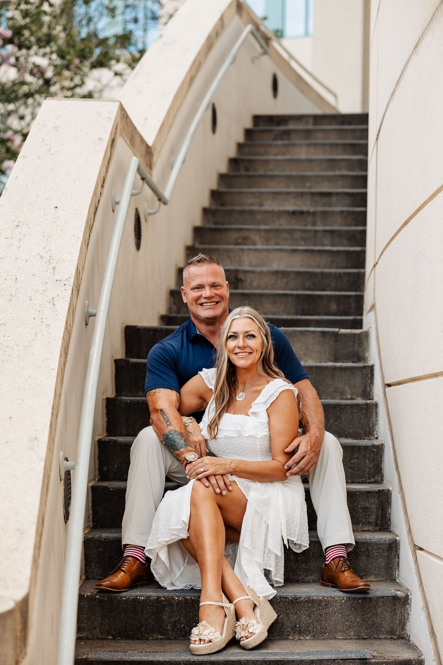 Engaged couple sits closely on outdoor stone steps, smiling warmly during a sunny urban engagement photoshoot.