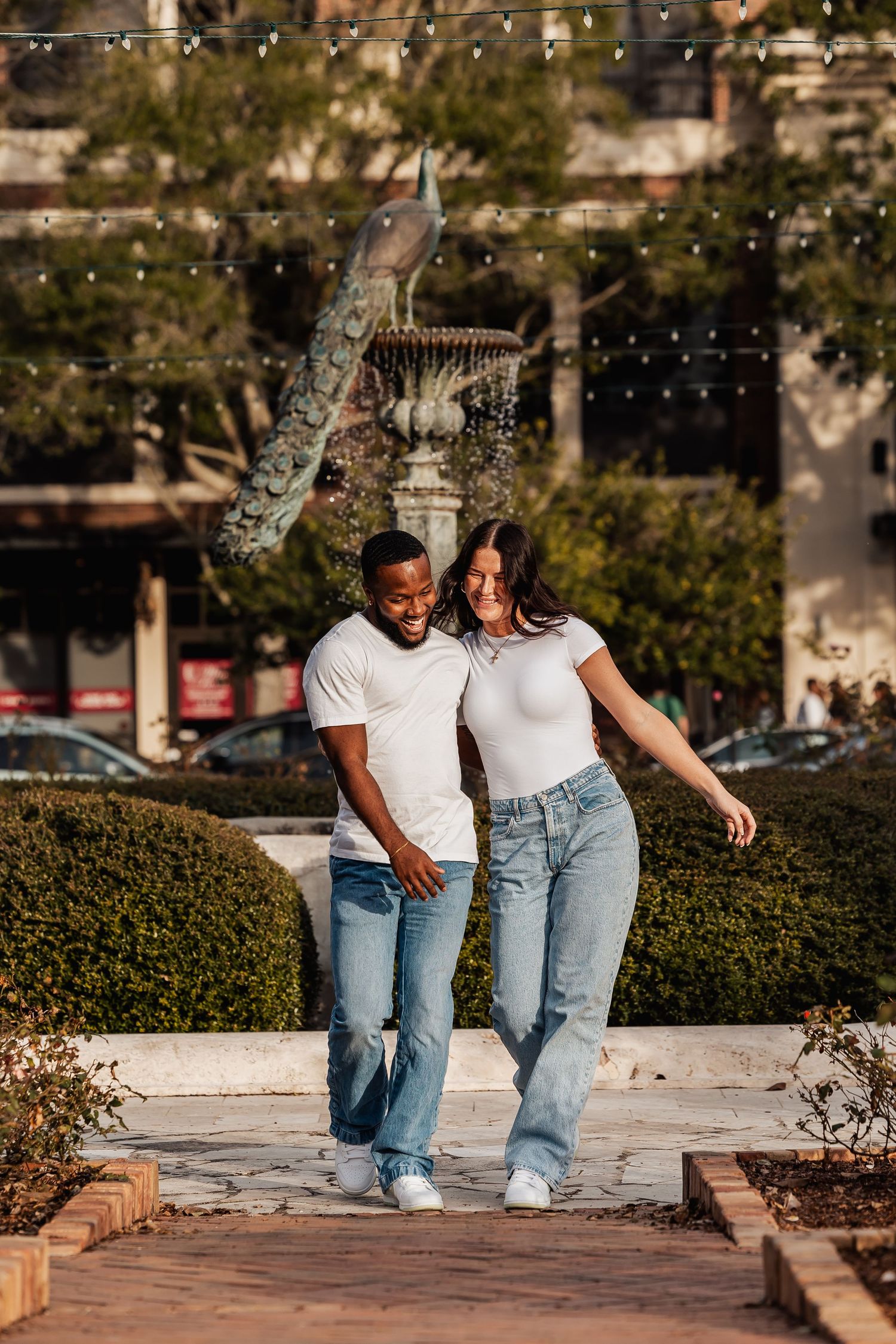 Stylish couple in matching white tees and jeans walking together near decorative fountain in park.