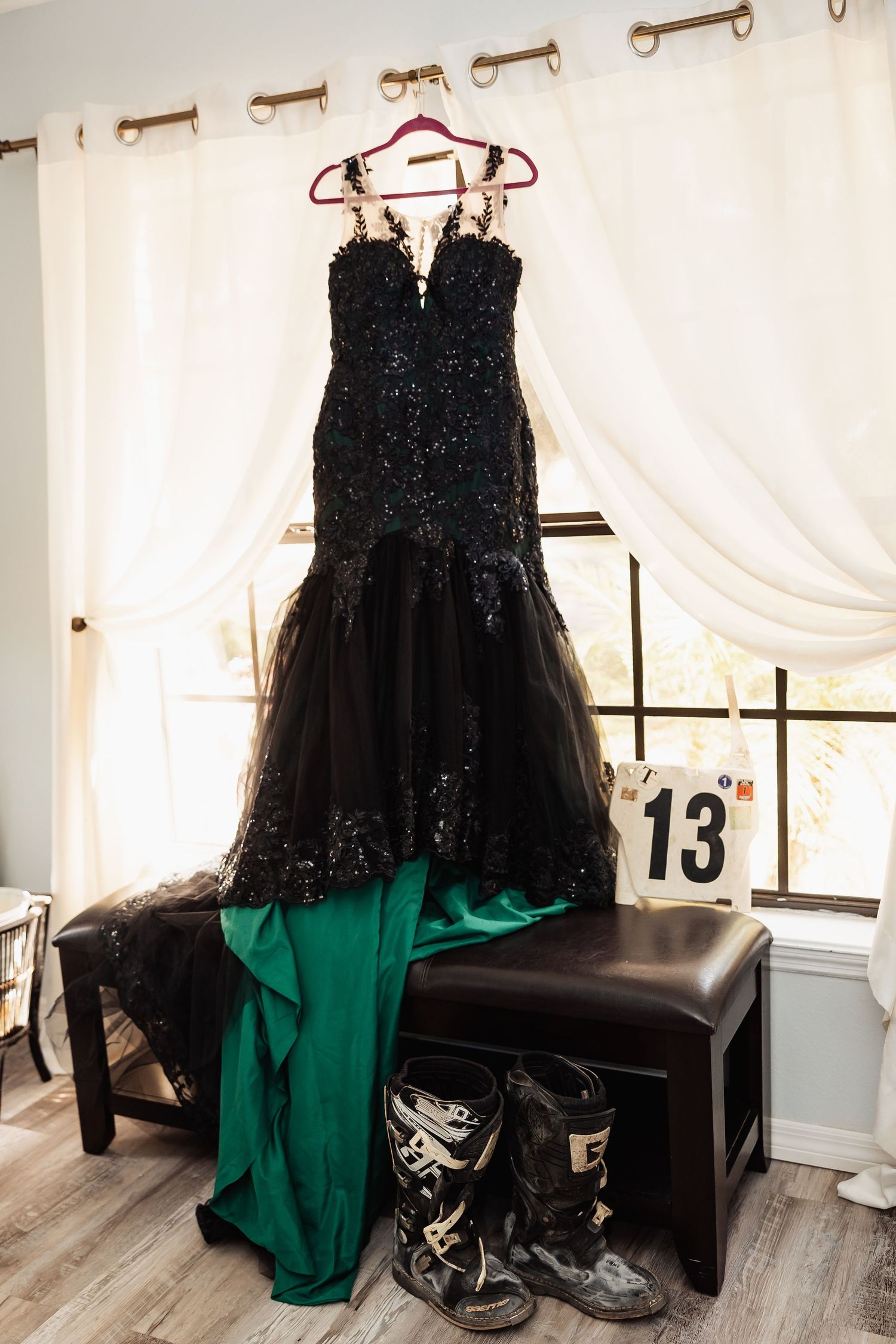 A black sequined mermaid wedding gown hanging by a window with motocross boots below it.