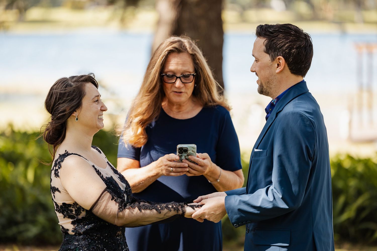 Bride and groom exchange vows with female officiant reading from phone during intimate outdoor wedding ceremony.