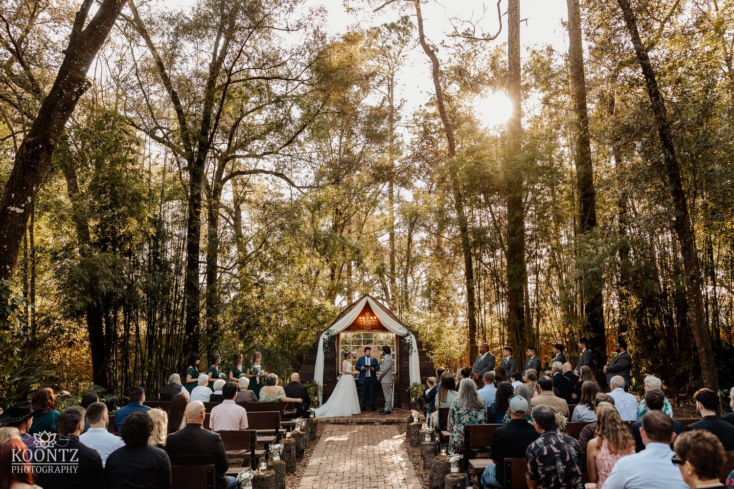 Outdoor woodland wedding ceremony with guests seated on wooden pews facing a rustic chapel altar.
