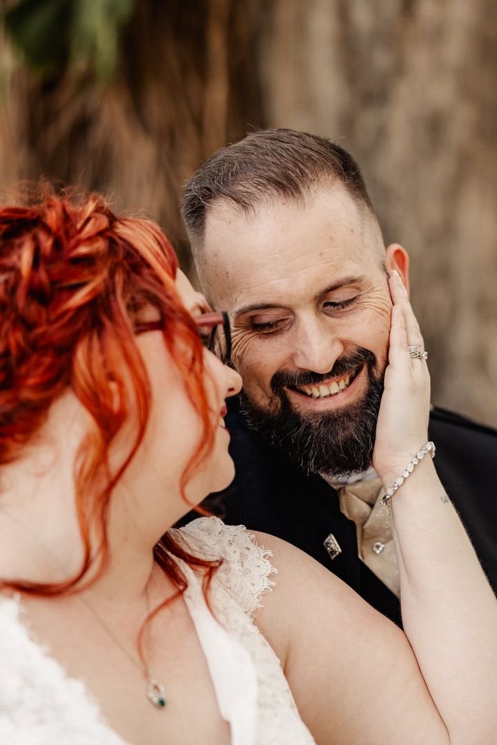 Bearded groom smiling joyfully as red-haired bride tenderly touches his face.