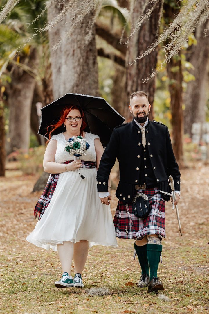Smiling bride with red hair and groom in Scottish kilt walk hand-in-hand under black umbrella among trees.