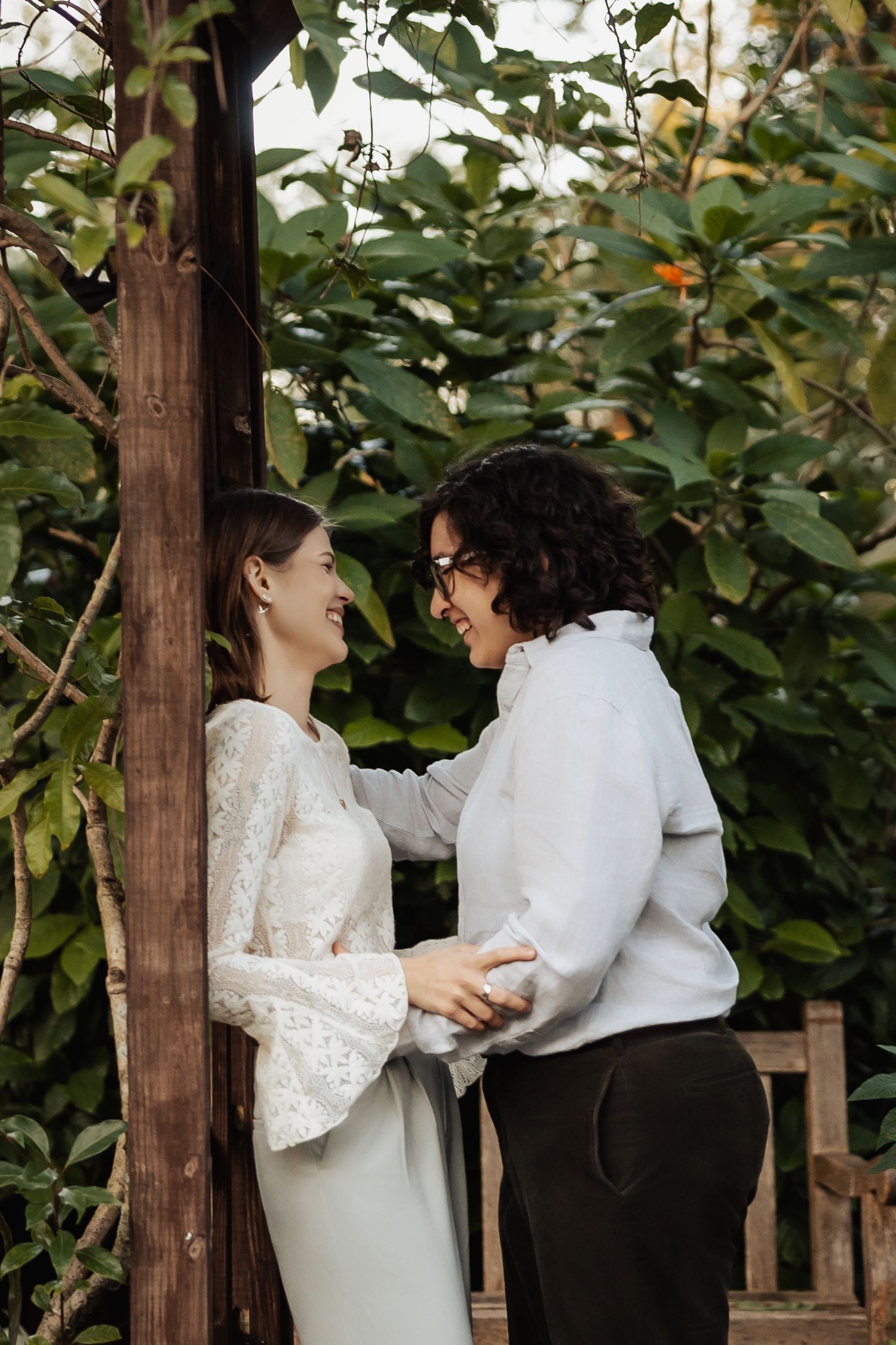 Two women laugh and embrace near a wooden garden arbor surrounded by lush green foliage.