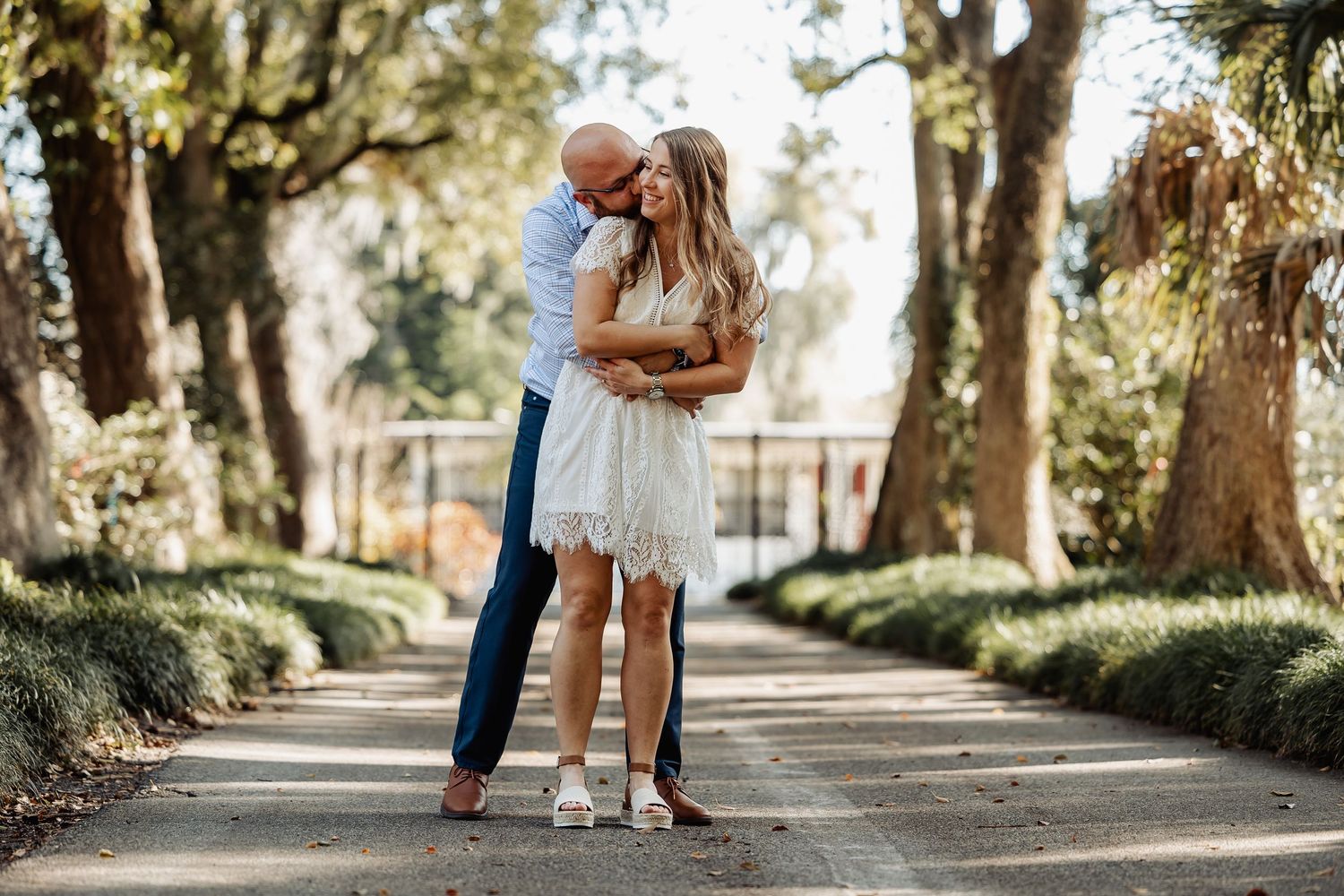 Man embracing woman in white lace dress on tree-lined garden pathway in warm sunlight.