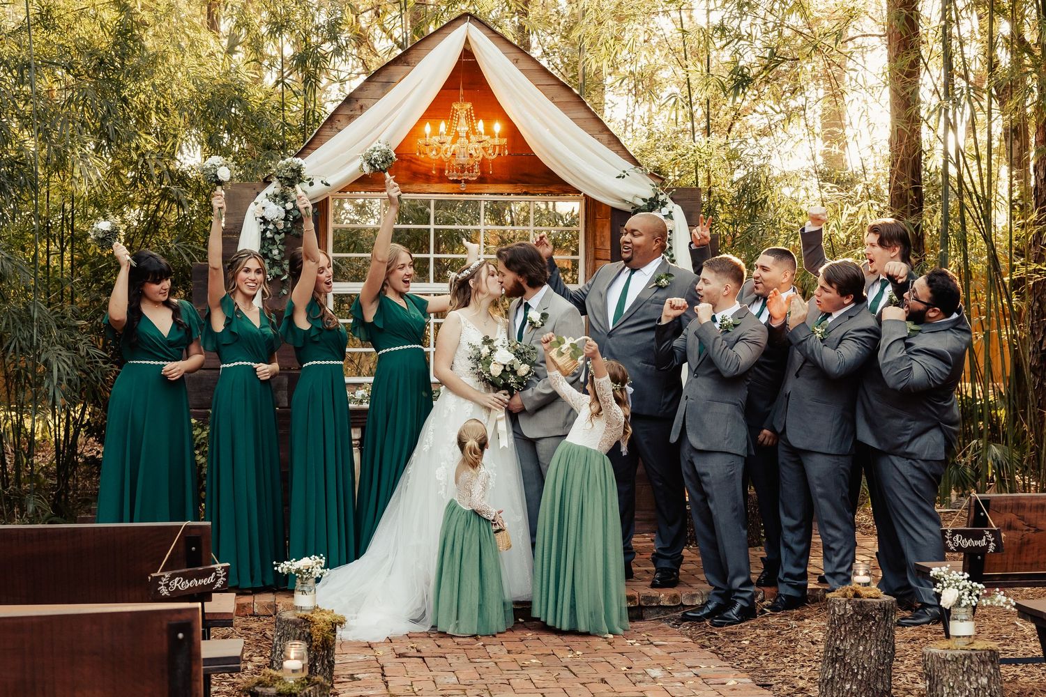 Wedding party celebrates with joyful poses outside rustic chapel surrounded by trees.