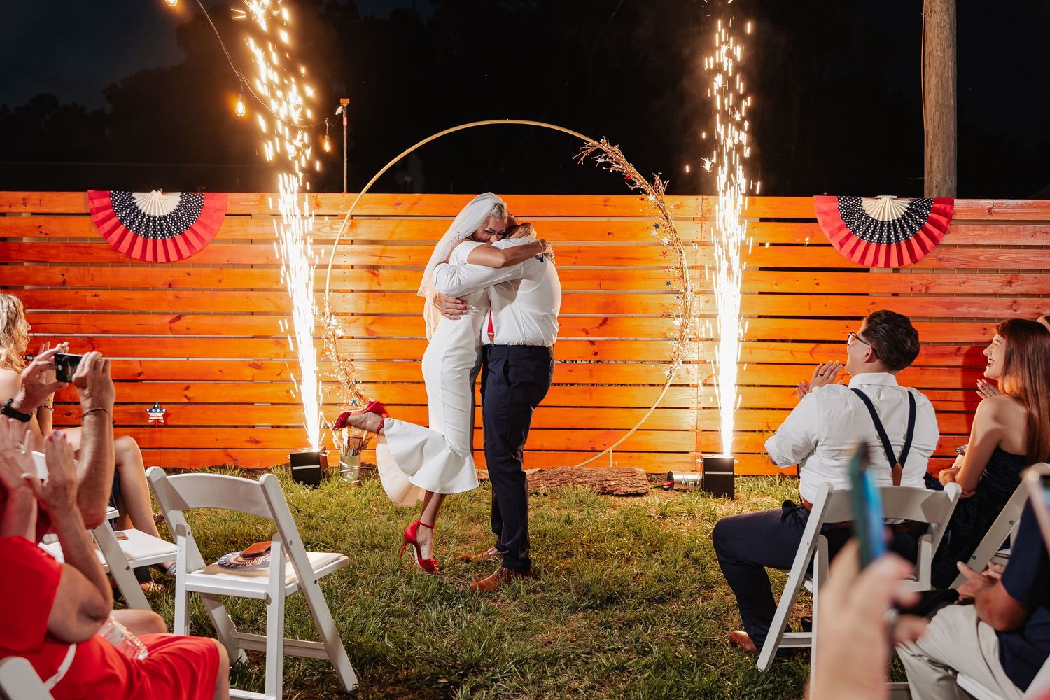 Newlyweds kiss under circular arch with sparkler fountains as guests cheer at outdoor patriotic wedding ceremony.
