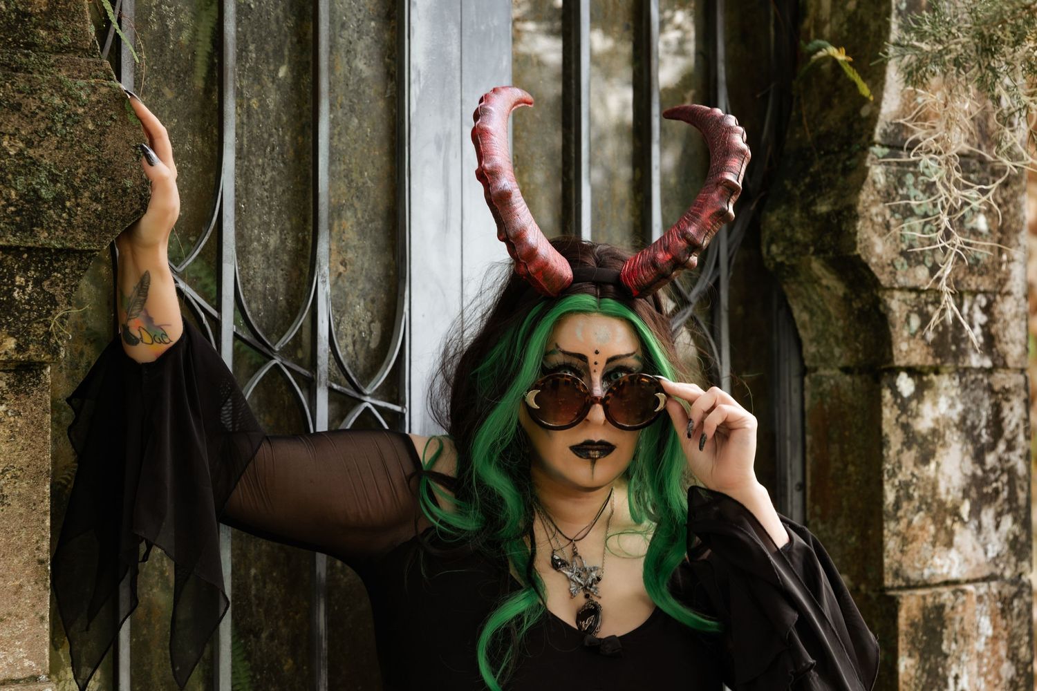 A woman with green hair and red demon horns wears round sunglasses posing against an iron gate.