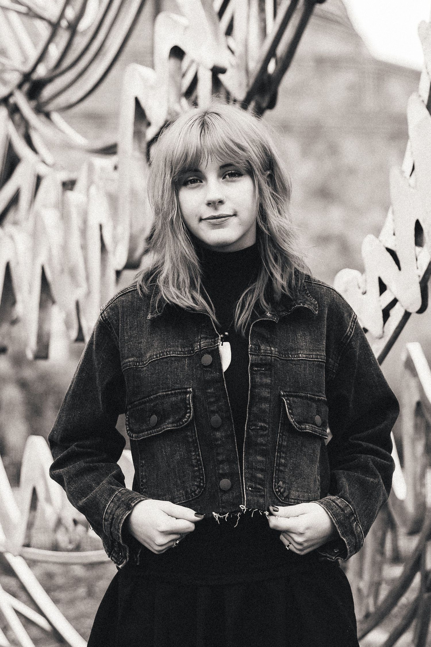 Black and white portrait of a young blonde girl in a denim jacket smiling outdoors near a metal sculpture.