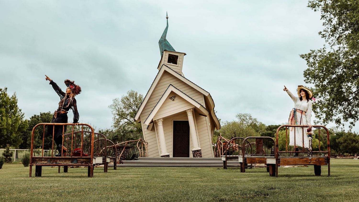 Two costumed figures pose dramatically on iron bed frames flanking a white country chapel.
