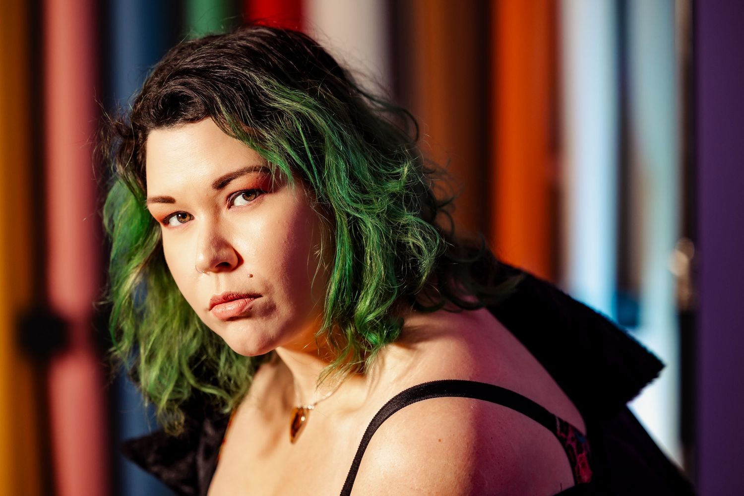 Young woman with green-tipped dark hair wearing black top poses against colorful blurred background.