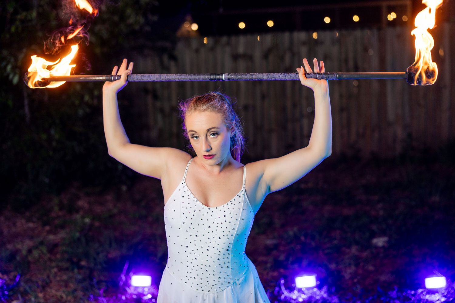 Female performer holding a double-ended flaming staff overhead in white dress during nighttime show.