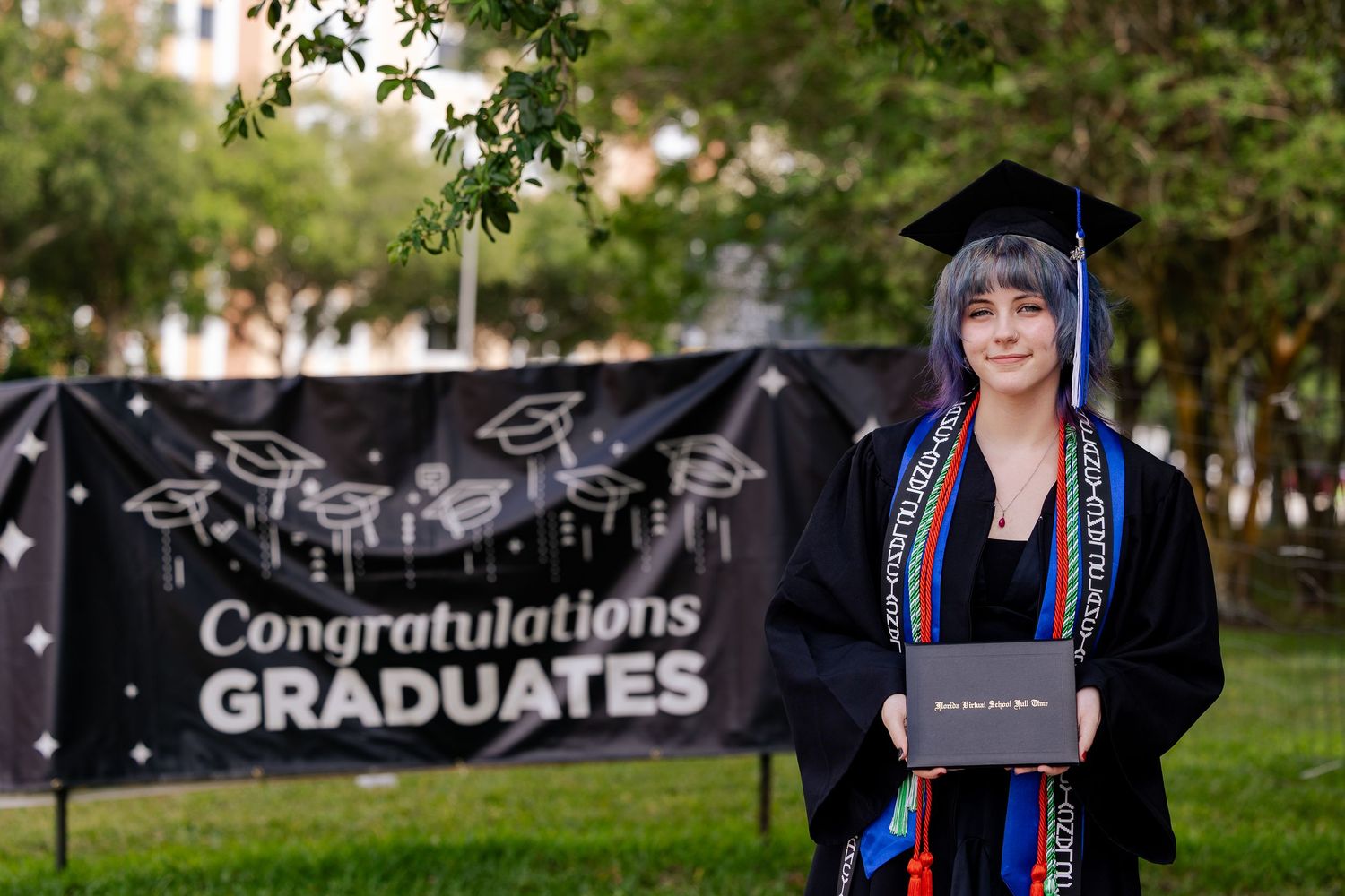 Graduate in cap and gown holds diploma in front of Congratulations Graduates banner outdoors.