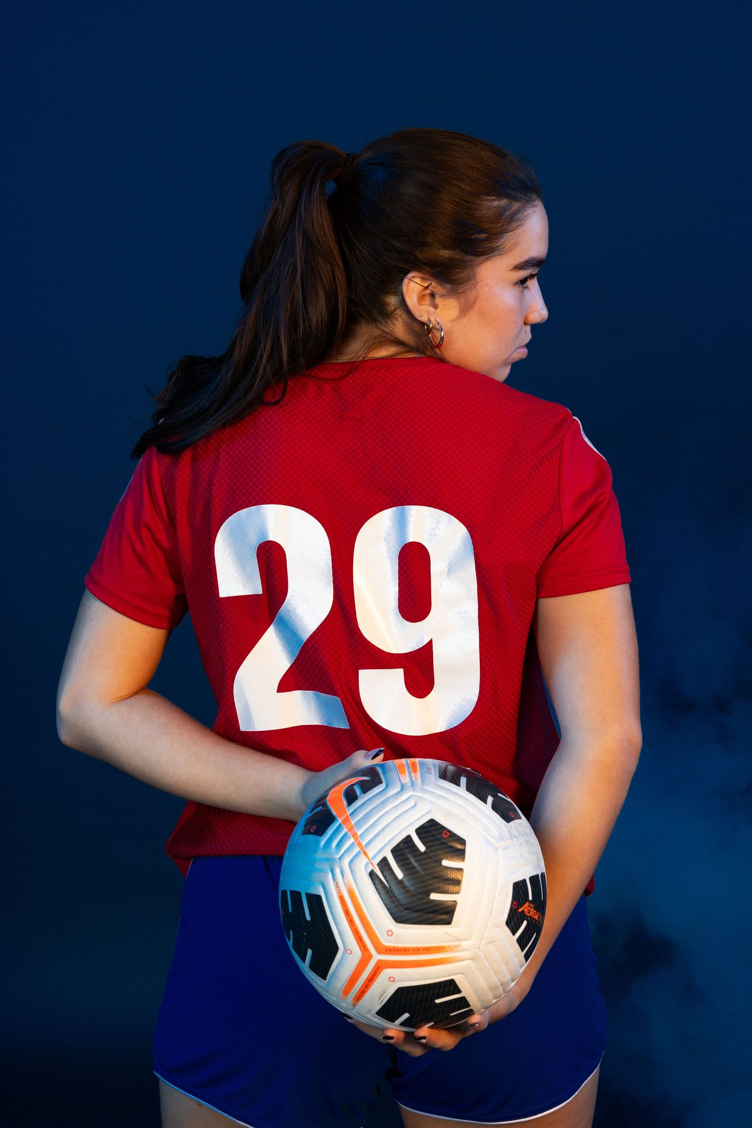 Female soccer player wearing red jersey number 29 holding a soccer ball, viewed from behind against dark blue background.