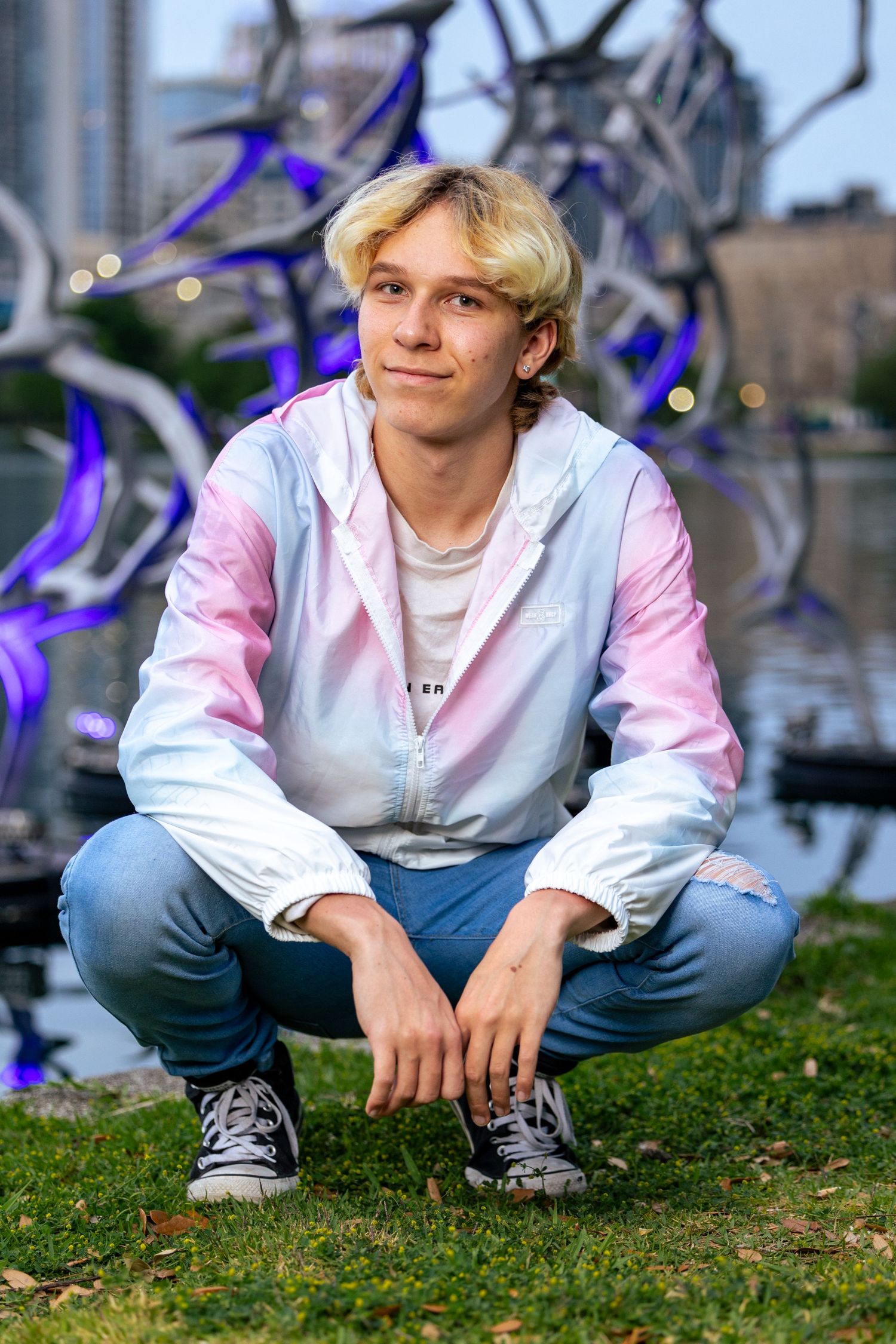 Teen boy with blonde hair crouching on grass, wearing a pink and white jacket and jeans, with a colorful sculpture behind him.