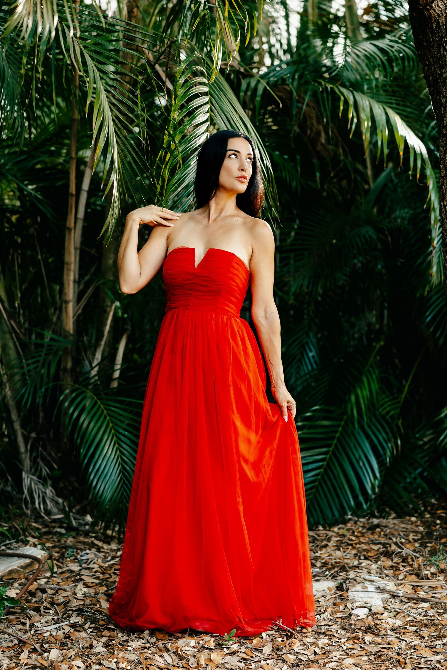 Woman in strapless red chiffon gown poses among lush tropical palm trees in an elegant outdoor fashion portrait.