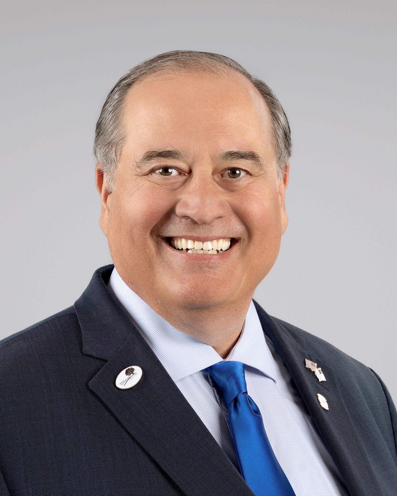 Professional headshot of a smiling man in a dark suit with a blue tie and lapel pins.