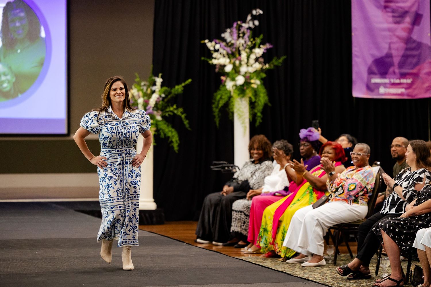Woman walks runway in blue floral dress at fashion show with seated audience.