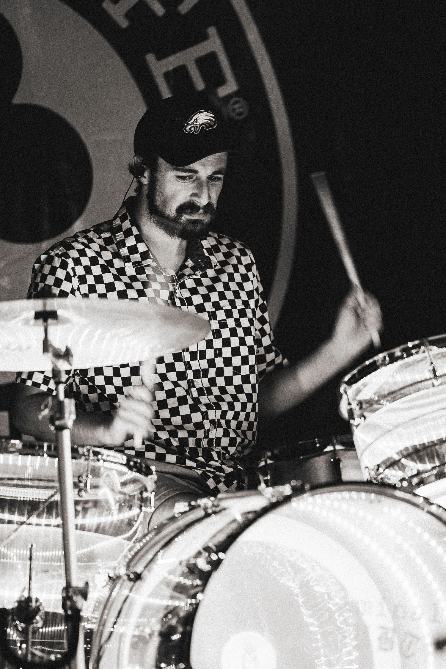 A bearded drummer in a checkered shirt plays drums in a black and white concert photo.