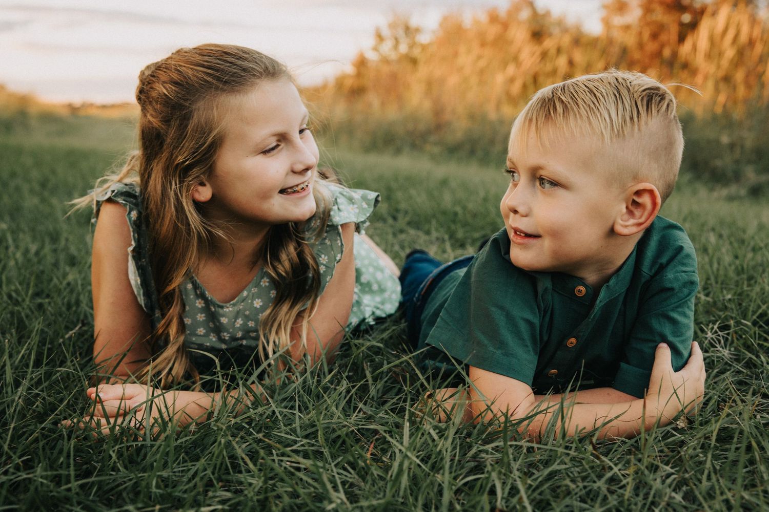 Young girl and boy lying in grass at golden hour, smiling and looking at each other.