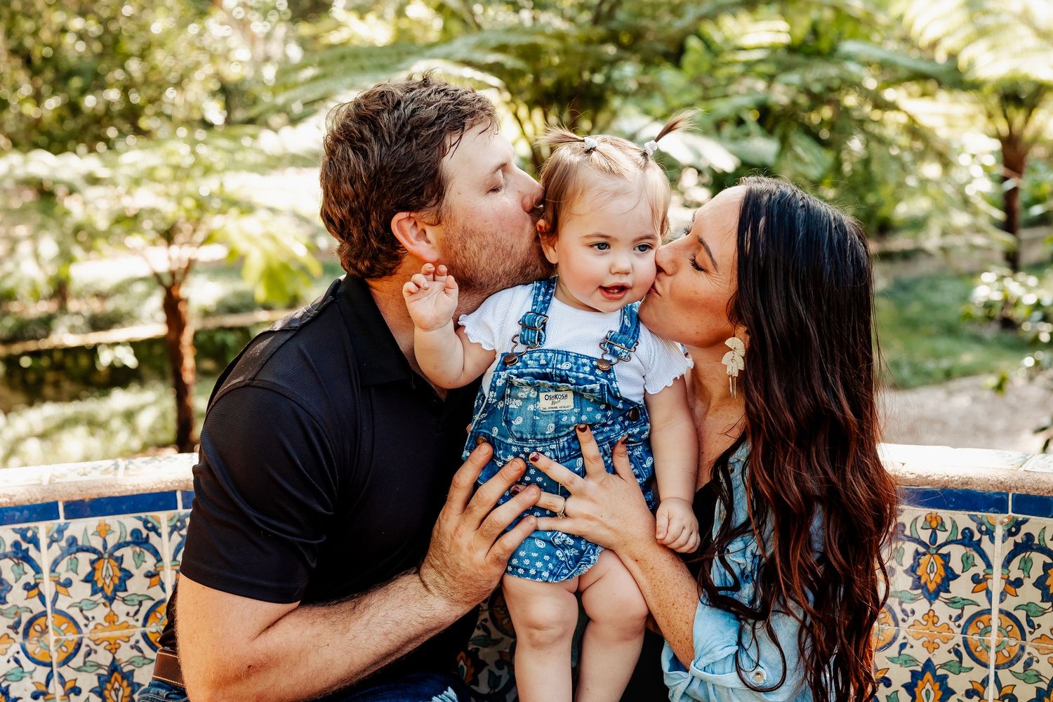 Parents kiss their smiling baby girl on the cheeks while seated outdoors in a lush green garden.