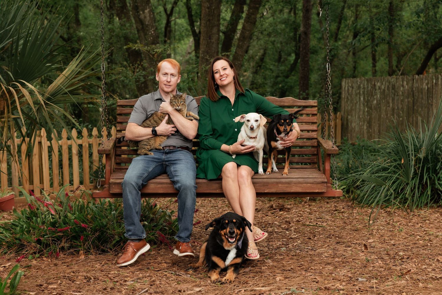A couple sits on a wooden bench outdoors with three dogs in a garden setting.