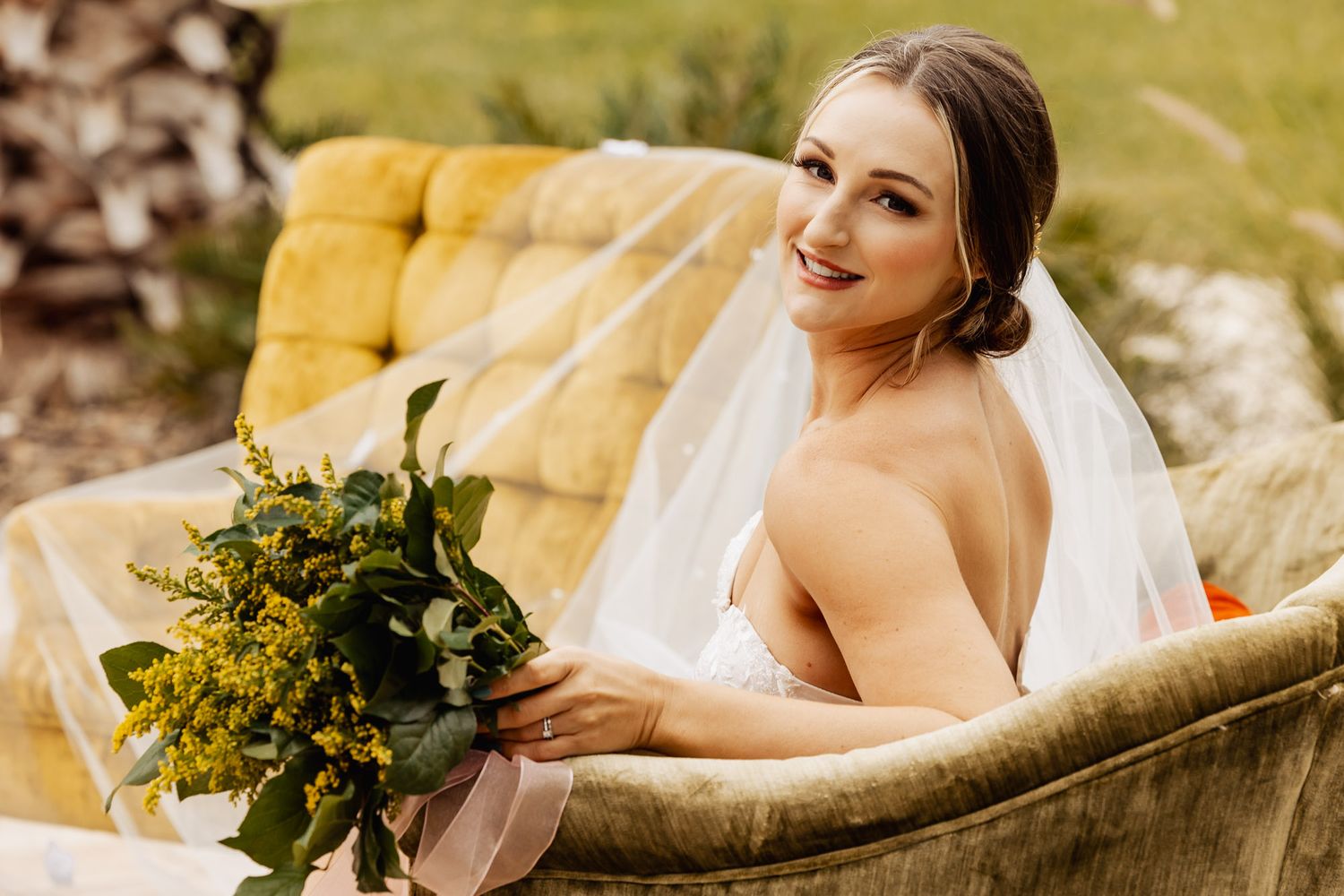 A bride wearing a white wedding dress sits on a yellow vintage couch outdoors while holding a green and yellow bouquet.