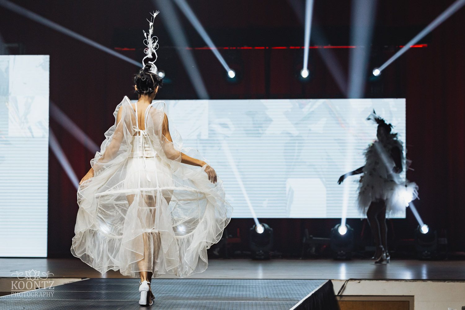 Models showcase elegant white flowing dresses and dramatic silhouettes on a sleek runway with dramatic blue lighting.