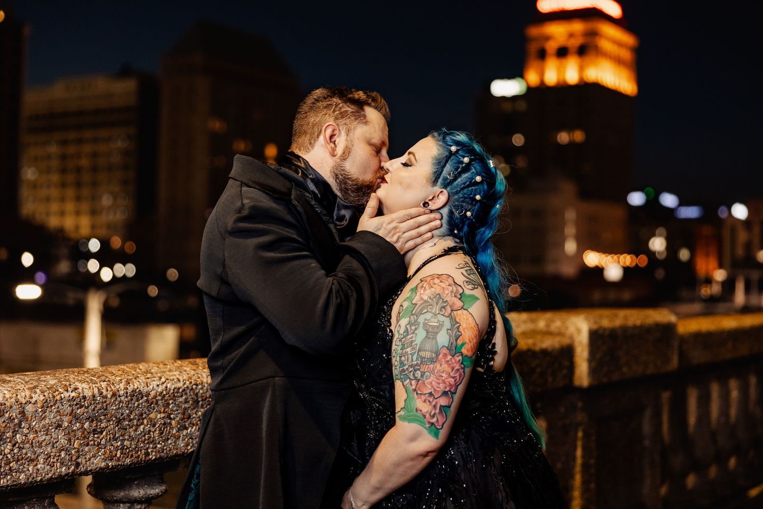 Gothic wedding couple kissing at night with illuminated city skyline and tower in background.