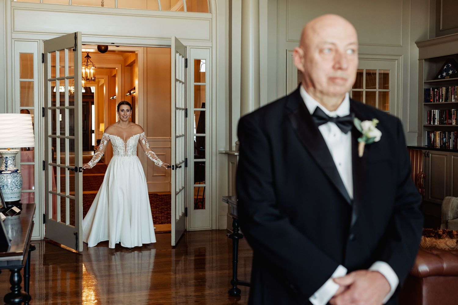 Bride in elegant long-sleeve lace gown stands in doorway as groom in tuxedo waits for first look.