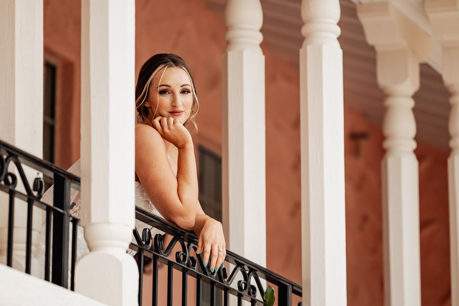 Blonde bride in white strapless gown leans gracefully on a balcony railing between white classical columns.