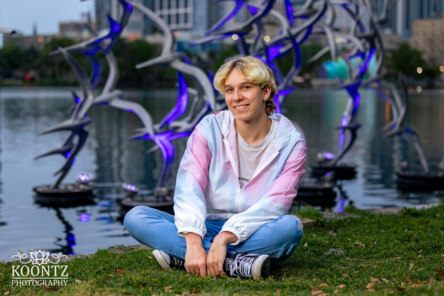 Outdoor portrait with artistic blue metal sculptures and water feature in background.