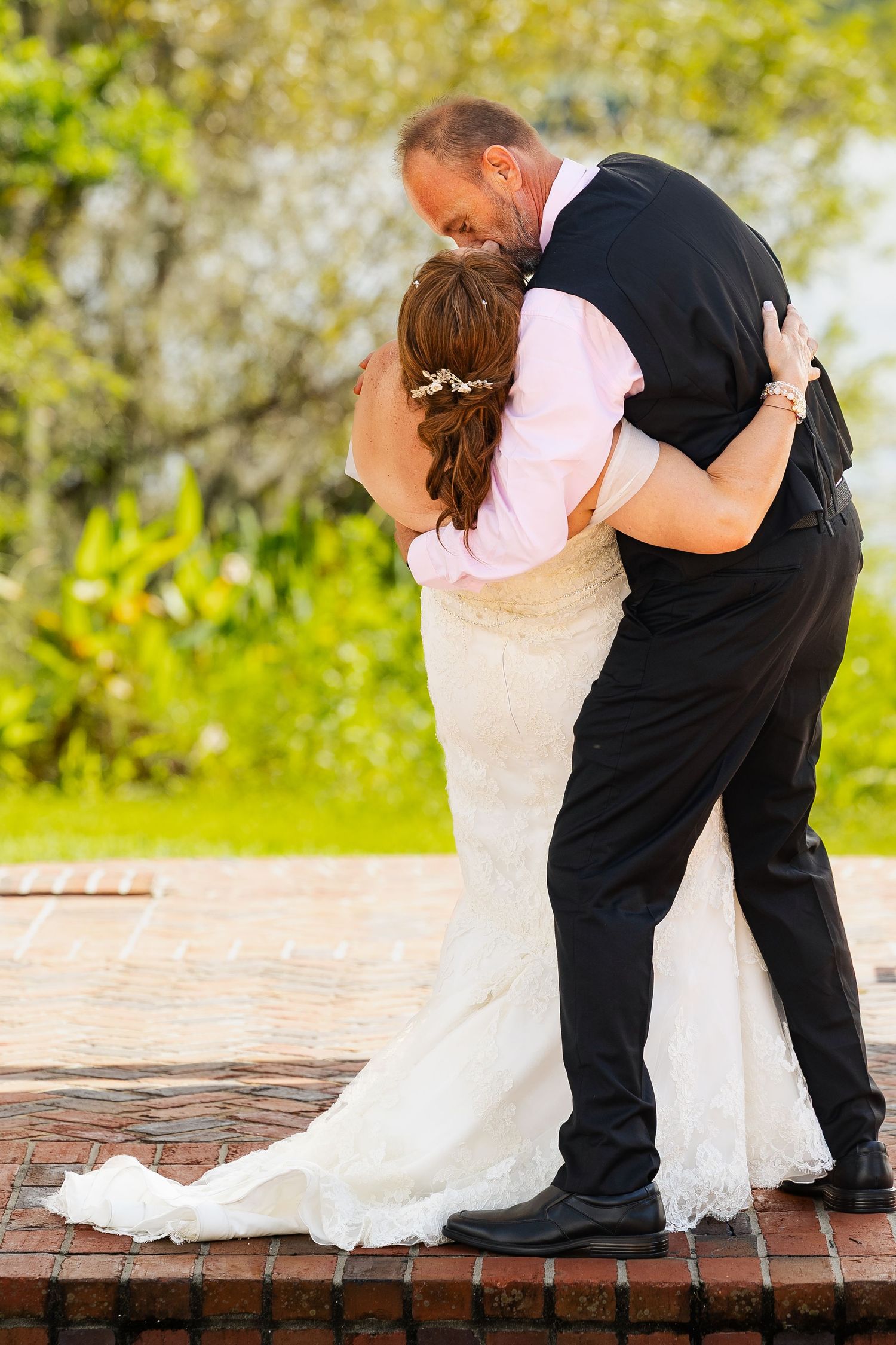 Bride and groom share a romantic first kiss embrace on brick terrace surrounded by lush greenery.