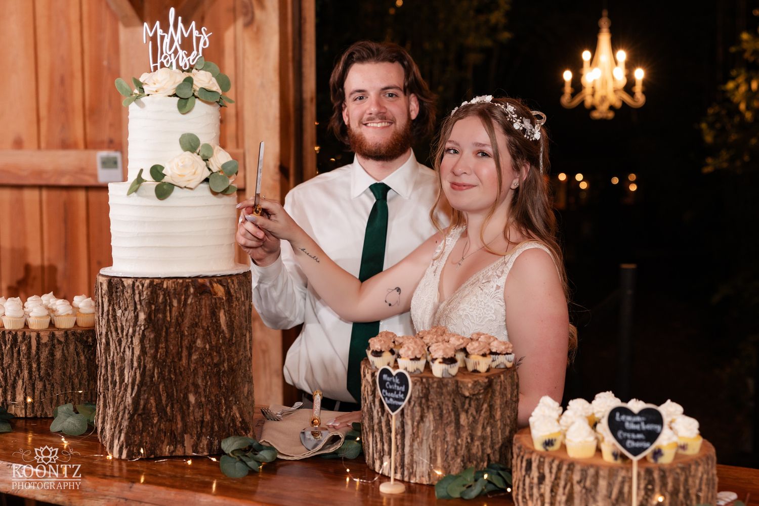 Bride and groom cut elegant white wedding cake displayed on wooden log stands at reception.