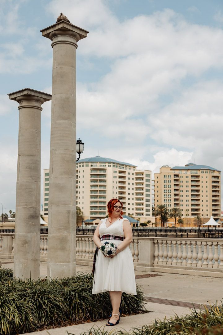 Bride in white tea-length dress holding bouquet beside stone columns with city skyline backdrop.