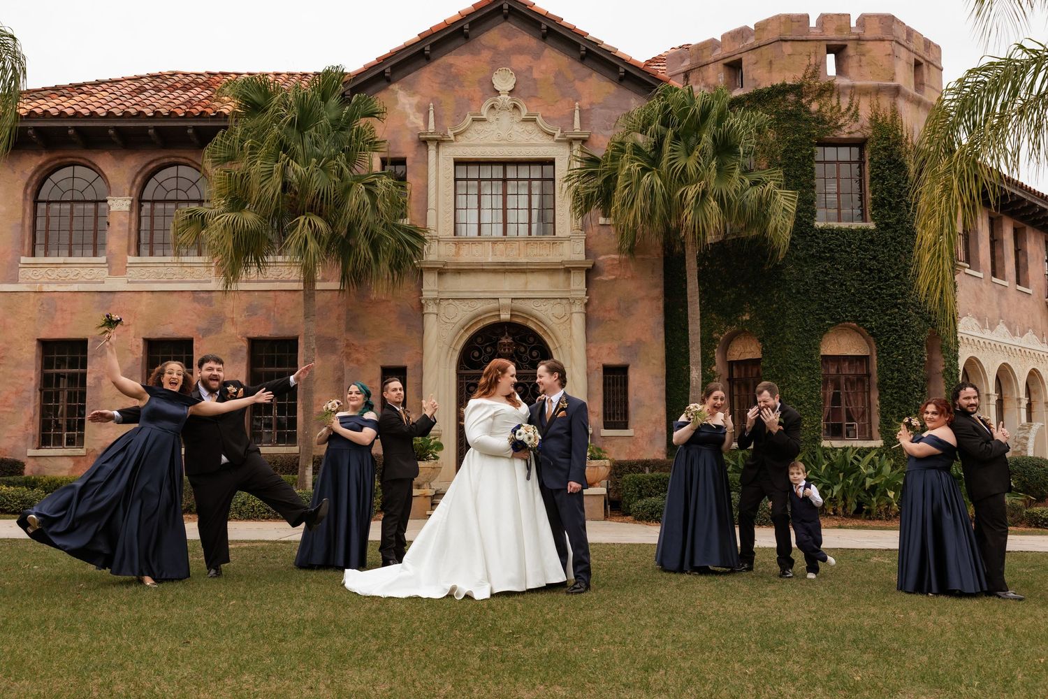 Wedding party celebrates outside a grand Spanish-style mansion with bridesmaids in navy gowns and groomsmen in black suits.