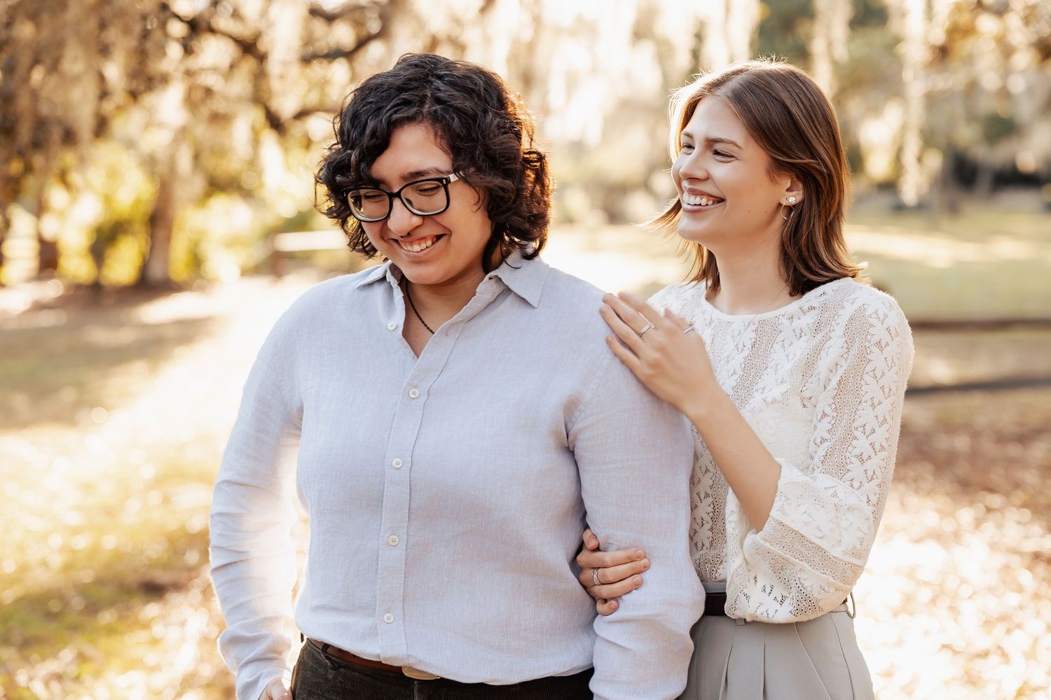 Two women laughing and embracing outdoors in a sunlit park during an engagement photo session.