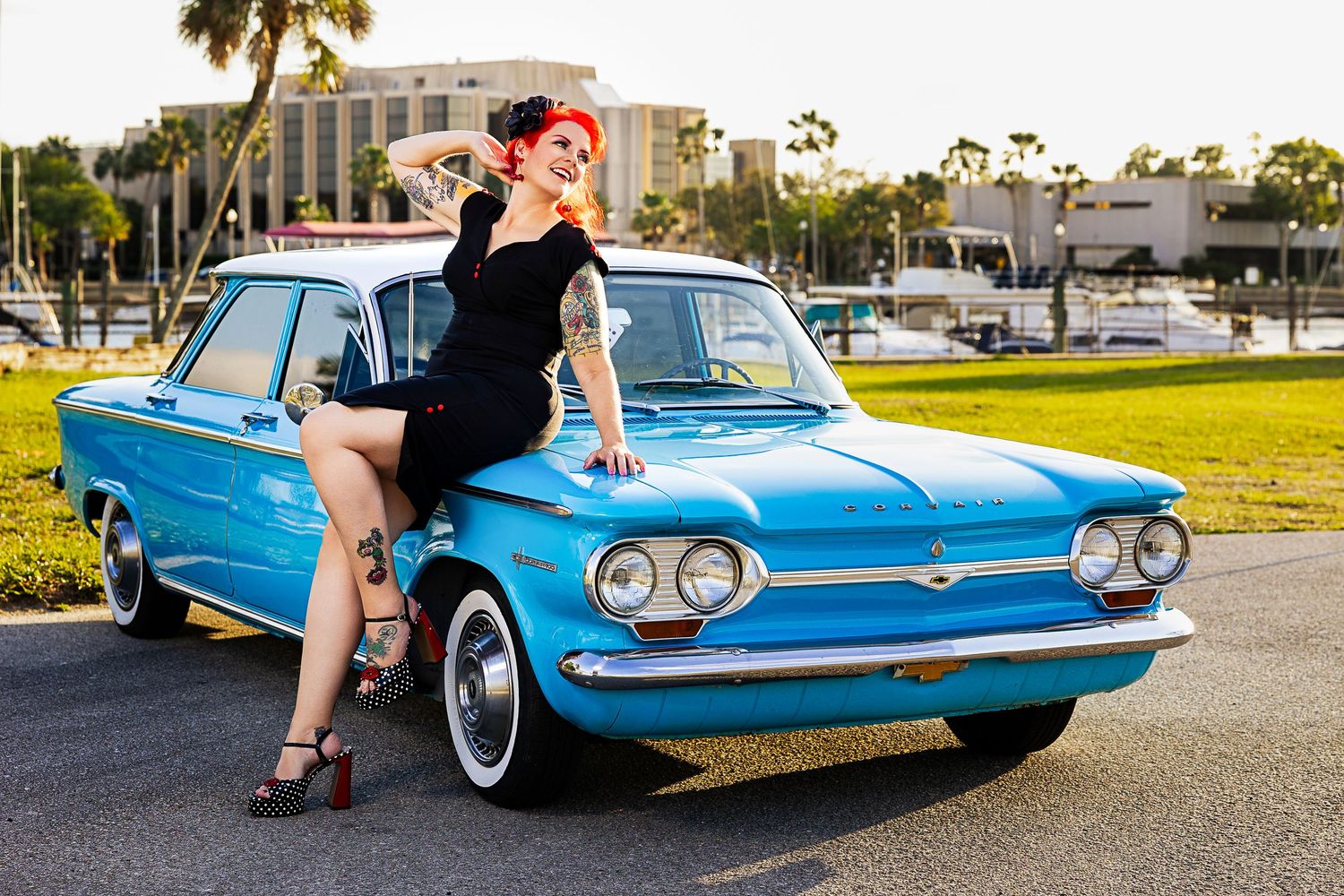 Tattooed pin-up woman in black dress posing with a classic blue Chevrolet Corvair on a sunny Florida day.