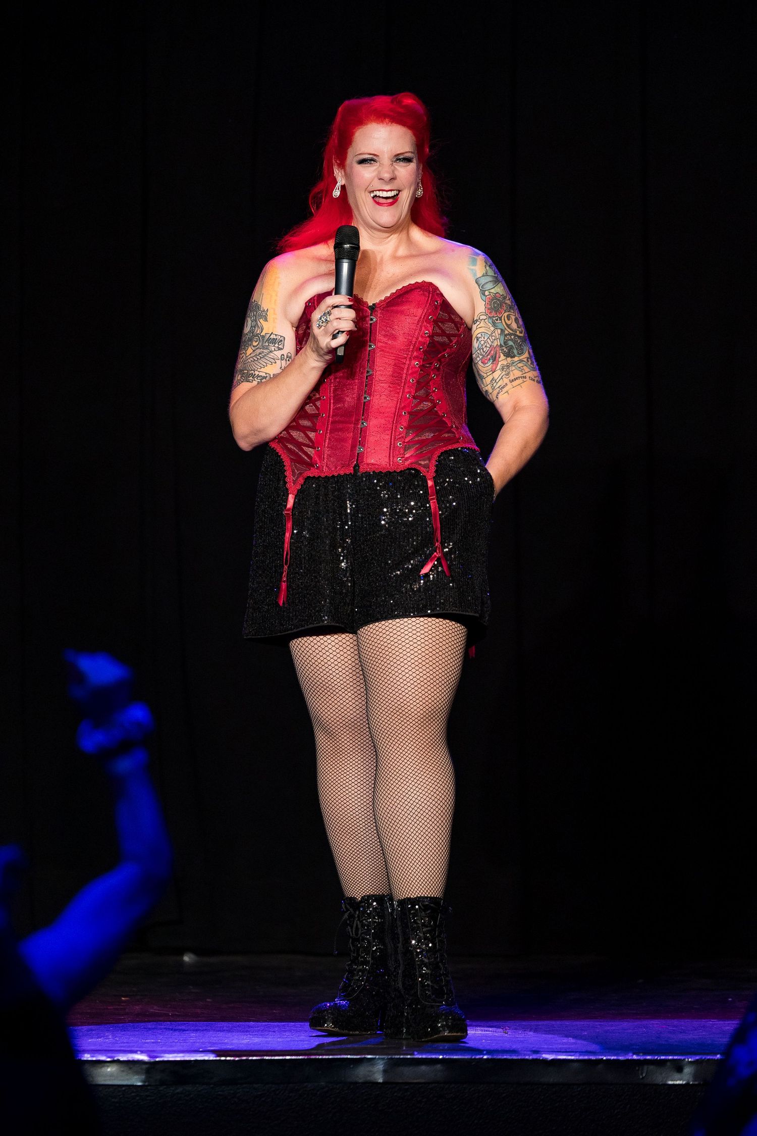 Plus-size performer in red corset and black sequin shorts holding microphone on stage.