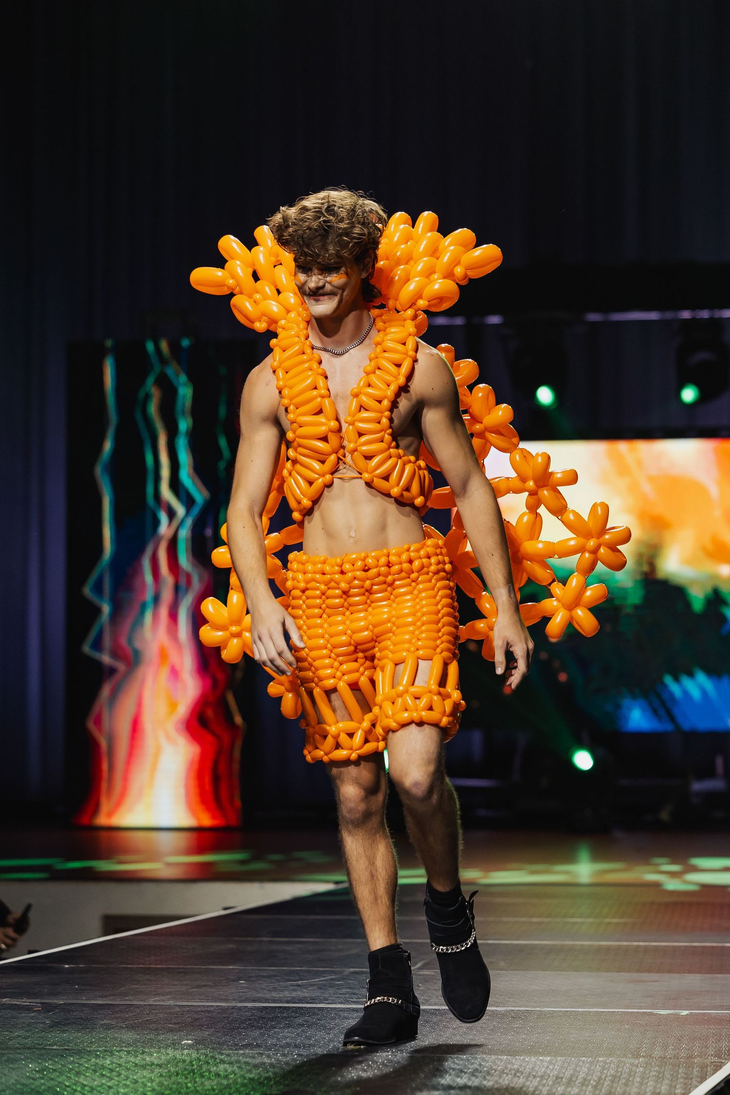 Male model walks runway wearing elaborate orange balloon art outfit with floral collar and shorts.