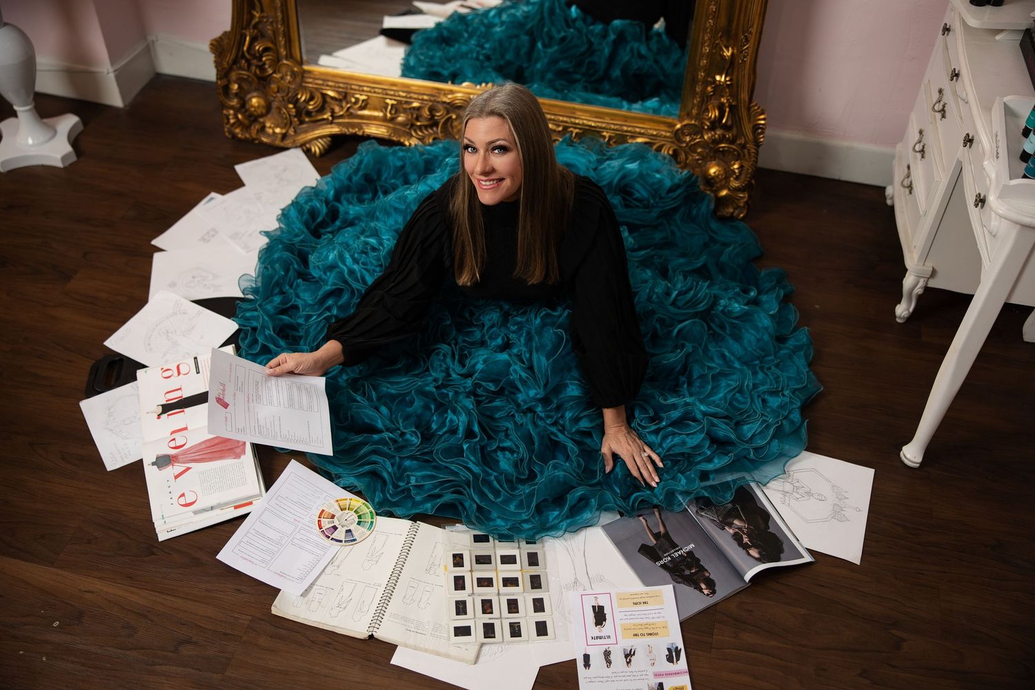 Smiling woman in teal ruffled skirt sitting on floor with fashion design documents in a boutique.