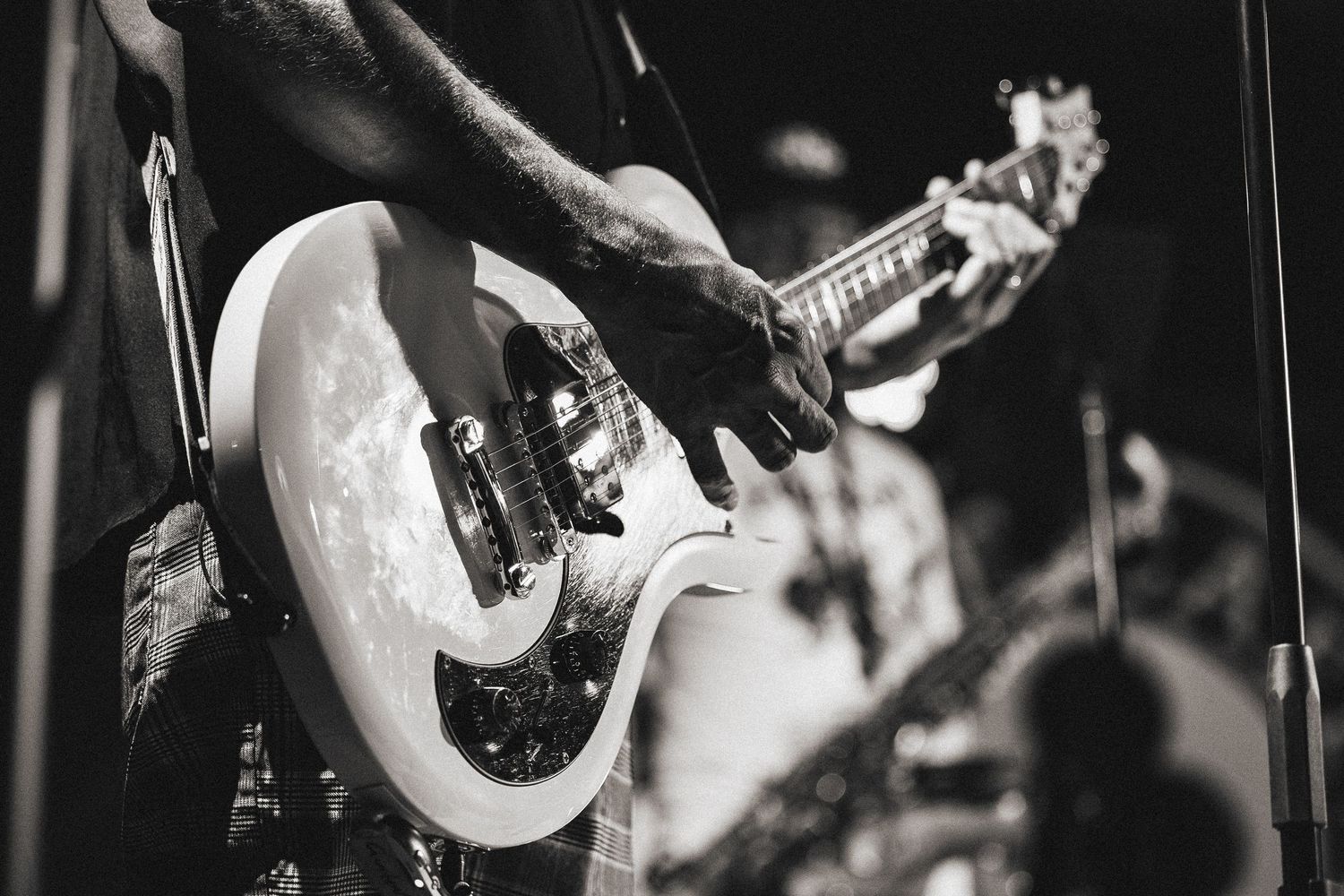 A close-up black and white shot of hands playing a white electric guitar during a live performance.